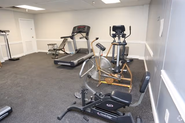 A small fitness room with exercise equipment including a treadmill, stationary bike, recumbent bike, and a weight bench with dumbbells. The room has gray carpeted flooring, white walls with a chair rail, and a white door in the background.