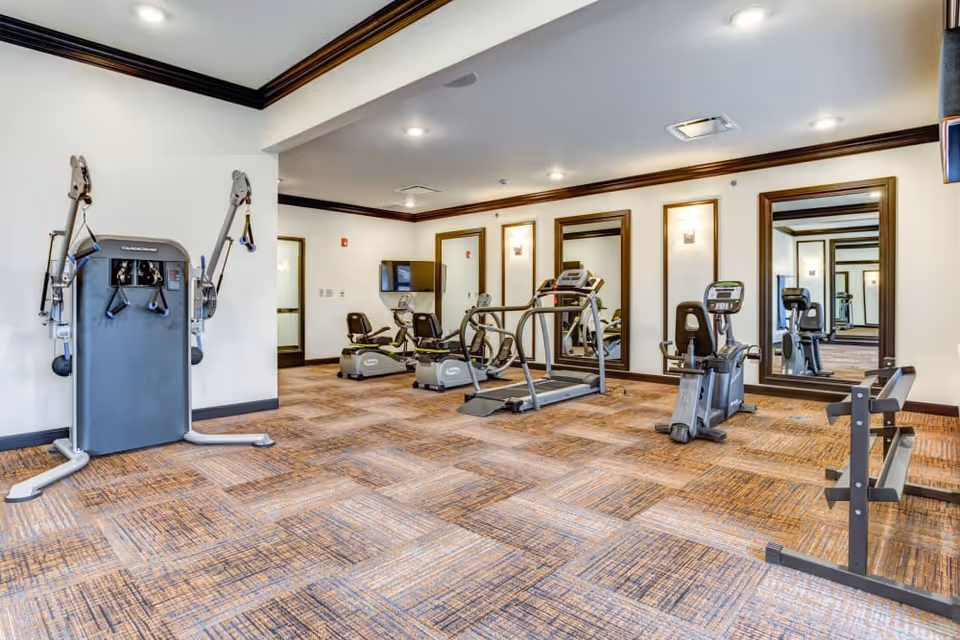 A fitness room with exercise equipment including a multi-function weight machine, two recumbent stationary bikes, a treadmill, and a weight bench. The room has large mirrors on the walls, recessed lighting, and carpeted flooring with a patterned design.