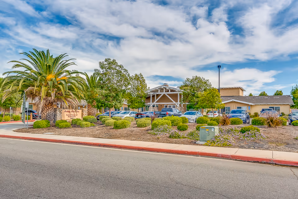 Exterior view of Sunny Rose Senior Living facility showing a single-story building with a parking lot in front, surrounded by trees, bushes, and a palm tree under a partly cloudy sky.