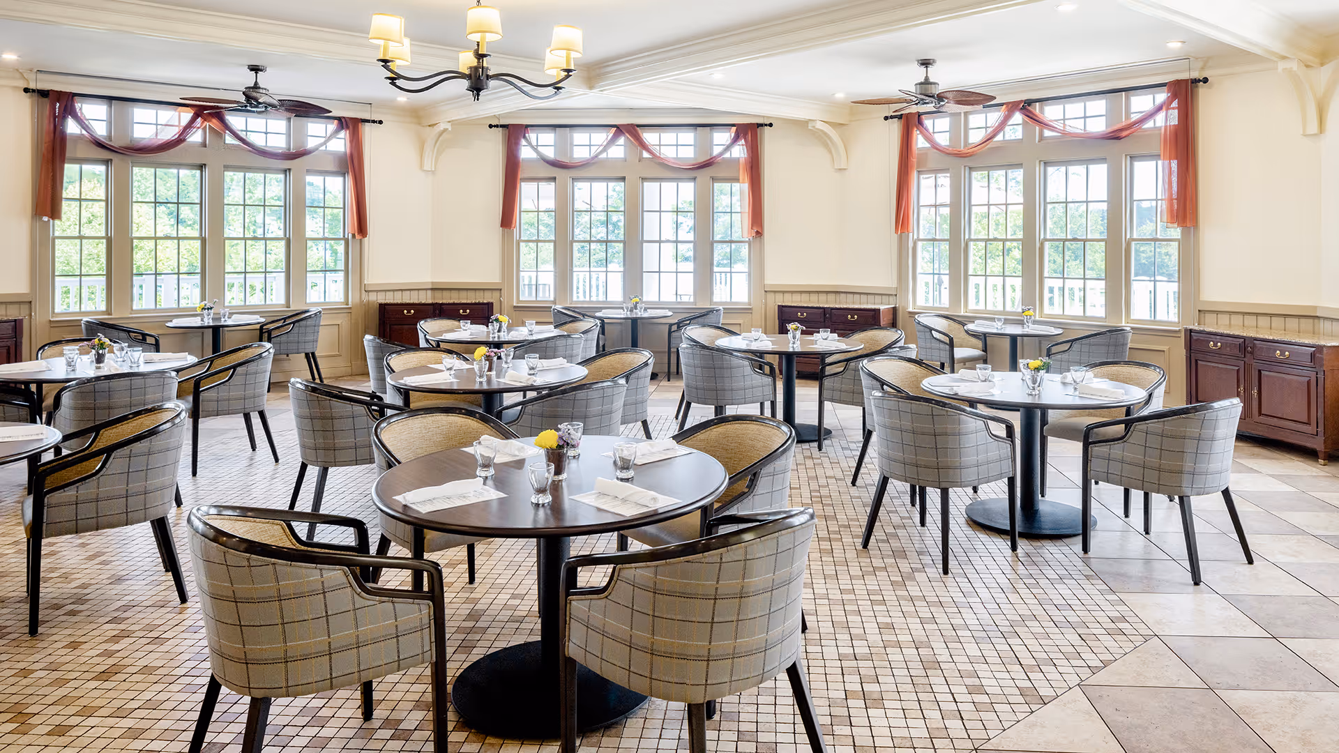 Bright dining room with multiple round tables set with glasses, napkins, and small flower arrangements. The room features large windows with red curtains, beige walls, tiled floors, and ceiling fans with lights.