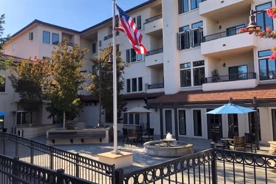 Courtyard with a flagpole, fountain, patio tables and umbrellas in front of a multi-story senior living building.