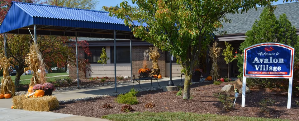Outdoor view of Avalon Village senior living facility entrance with a covered walkway, autumn decorations including pumpkins and hay bales, trees, and a welcome sign.