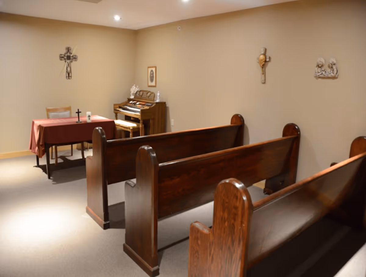 Interior of a small chapel room with wooden pews, a table covered with a red cloth holding two crosses, a wooden organ with a bench, and religious wall decorations including crosses and a framed picture.