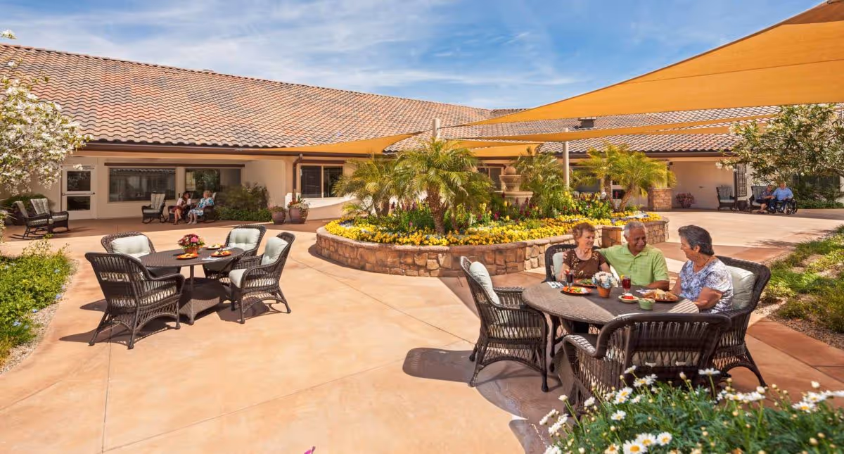 Outdoor courtyard area at Copper Creek Inn with several wicker tables and chairs. Three elderly people are seated at one table enjoying a meal and conversation. The courtyard features a central raised flower bed with palm trees and colorful flowers, shaded by large yellow fabric canopies. The building with a tiled roof surrounds the courtyard, and additional seating is visible near the building.