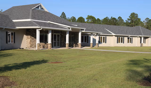 Single-story senior living building with a covered porch, stone columns, and a green lawn under a clear sky.