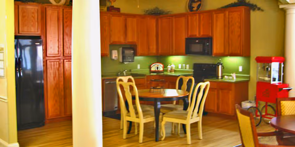 Interior view of a kitchen and dining area with wooden cabinets, a black refrigerator, a microwave, a dishwasher, and a round wooden table with four chairs. There is a red popcorn machine on the right side and green walls with under-cabinet lighting.