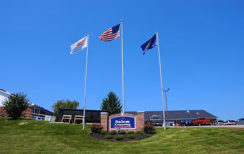 A grassy lawn with a brick 'Salem Crossing' sign, three flagpoles (one with an American flag) and the facility building under a clear blue sky.