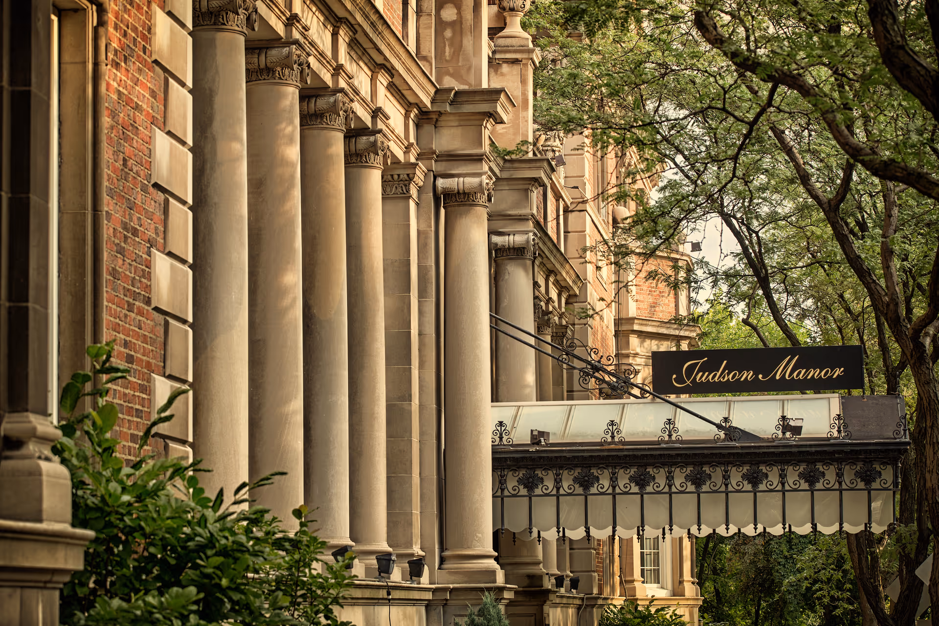 Exterior view of Judson Manor showing a row of large classical columns on a brick building with ornate architectural details. A black and gold sign reading 'Judson Manor' is mounted above a decorative awning. Trees and greenery surround the building.