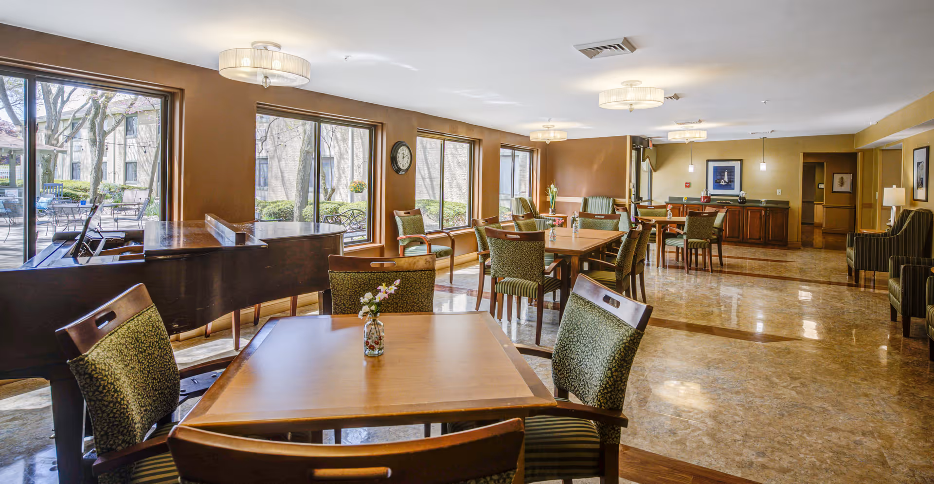 Bright senior community dining and lounge area with wooden tables and green upholstered chairs, a grand piano by large windows, and a serving counter in the back.