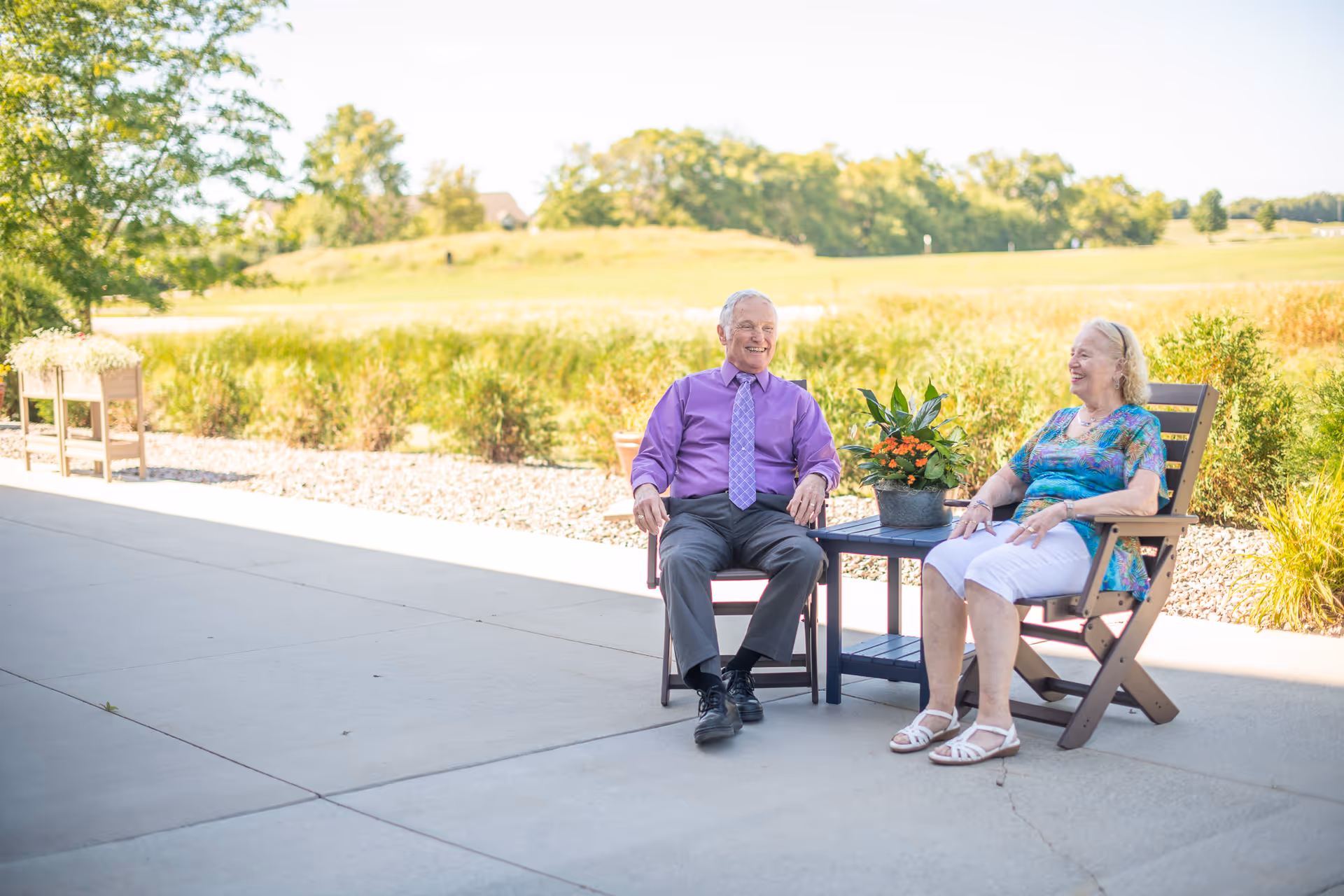 An elderly man and woman sitting outdoors on wooden chairs with a small table between them holding a potted plant, smiling and enjoying a sunny day with greenery and trees in the background.