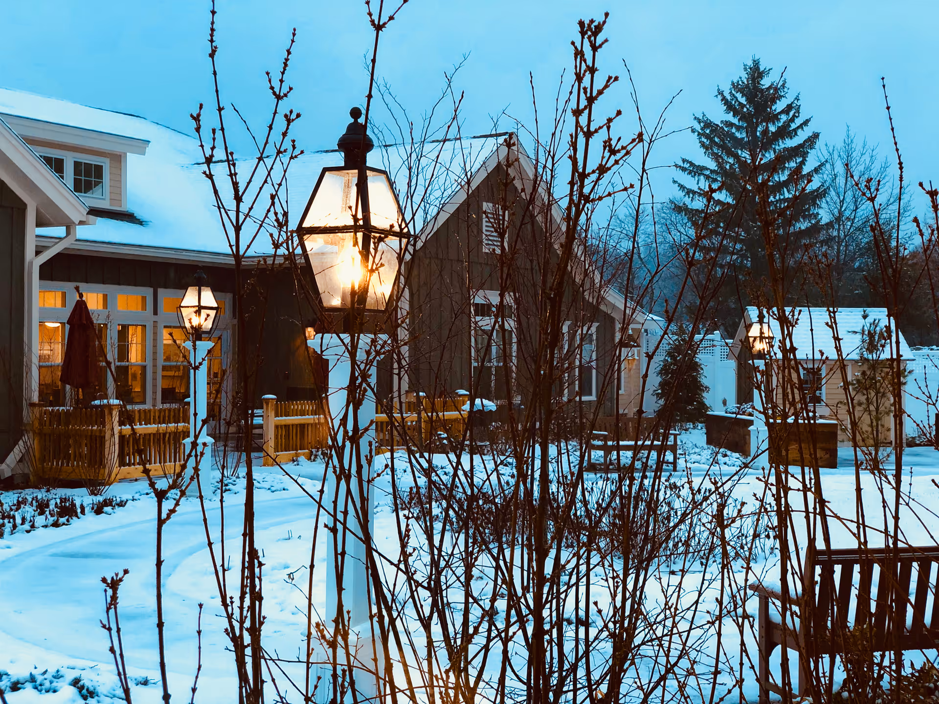 Snow-covered courtyard outside a building with lit lampposts, leafless shrubs, and a wooden fence in evening light.