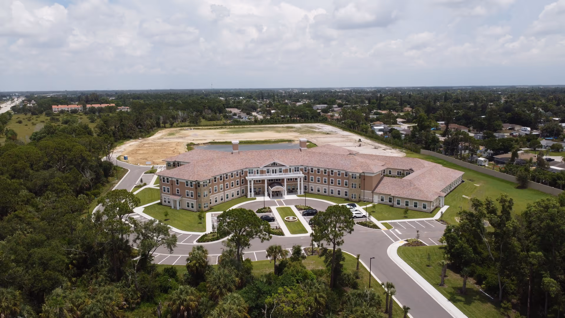 Aerial view of a large, three-story senior living facility building with a circular driveway and parking spaces in front, surrounded by green lawns and trees. The building has a tan exterior with white trim and a red-tiled roof. The surrounding area includes a mix of wooded land and residential neighborhoods under a partly cloudy sky.