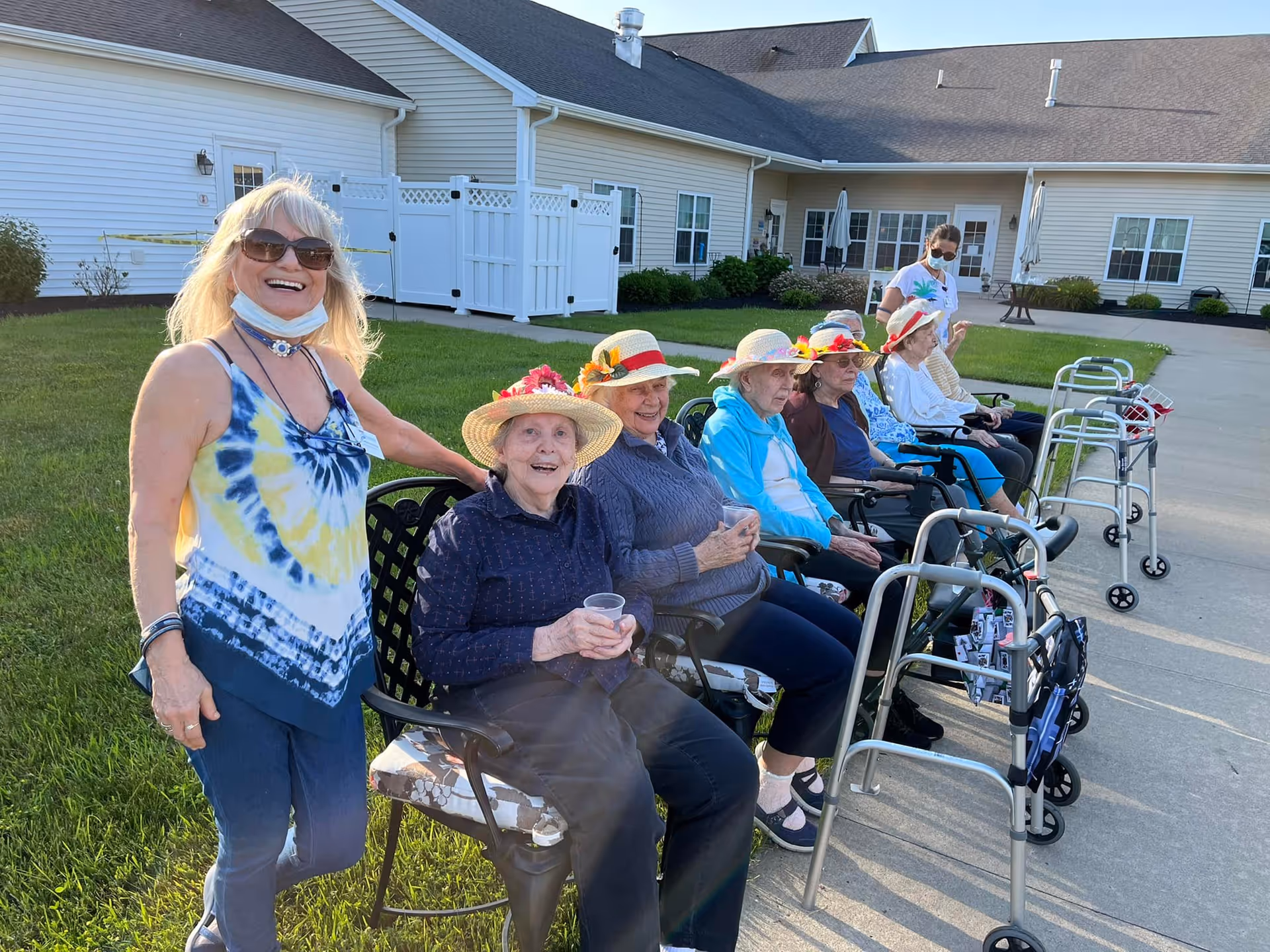 A group of elderly women sitting outside on chairs along a sidewalk, wearing colorful hats with flowers. A woman standing next to them is smiling and wearing sunglasses and a tie-dye tank top. Another person in the background is wearing a mask and standing near the building. Walkers are positioned in front of the seated women. The setting is a sunny day outside a light-colored building with grass and paved areas.