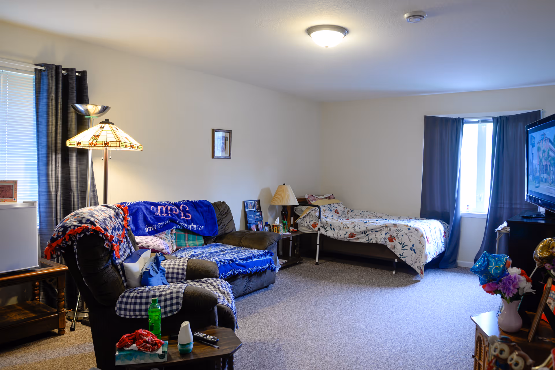 A cozy living space with a single bed covered in floral bedding near a window with dark curtains. There is a dark armchair and a loveseat with colorful blankets and pillows. A floor lamp with a stained glass shade stands beside the loveseat. A small wooden table holds a green bottle, a remote control, and other small items. A television is on a stand opposite the seating area, and the room has beige carpet and white walls.