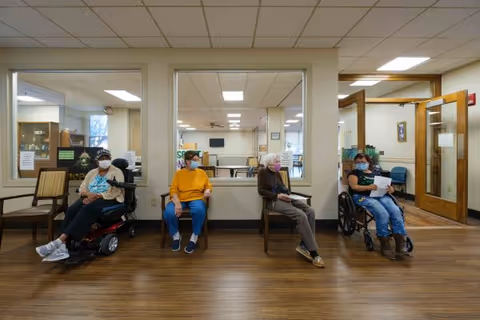 Four elderly individuals wearing face masks are seated in a waiting area inside a facility. Two are in wheelchairs and two are seated on chairs. The room has wooden flooring, large windows looking into another room, and a door to the right leading to another area.