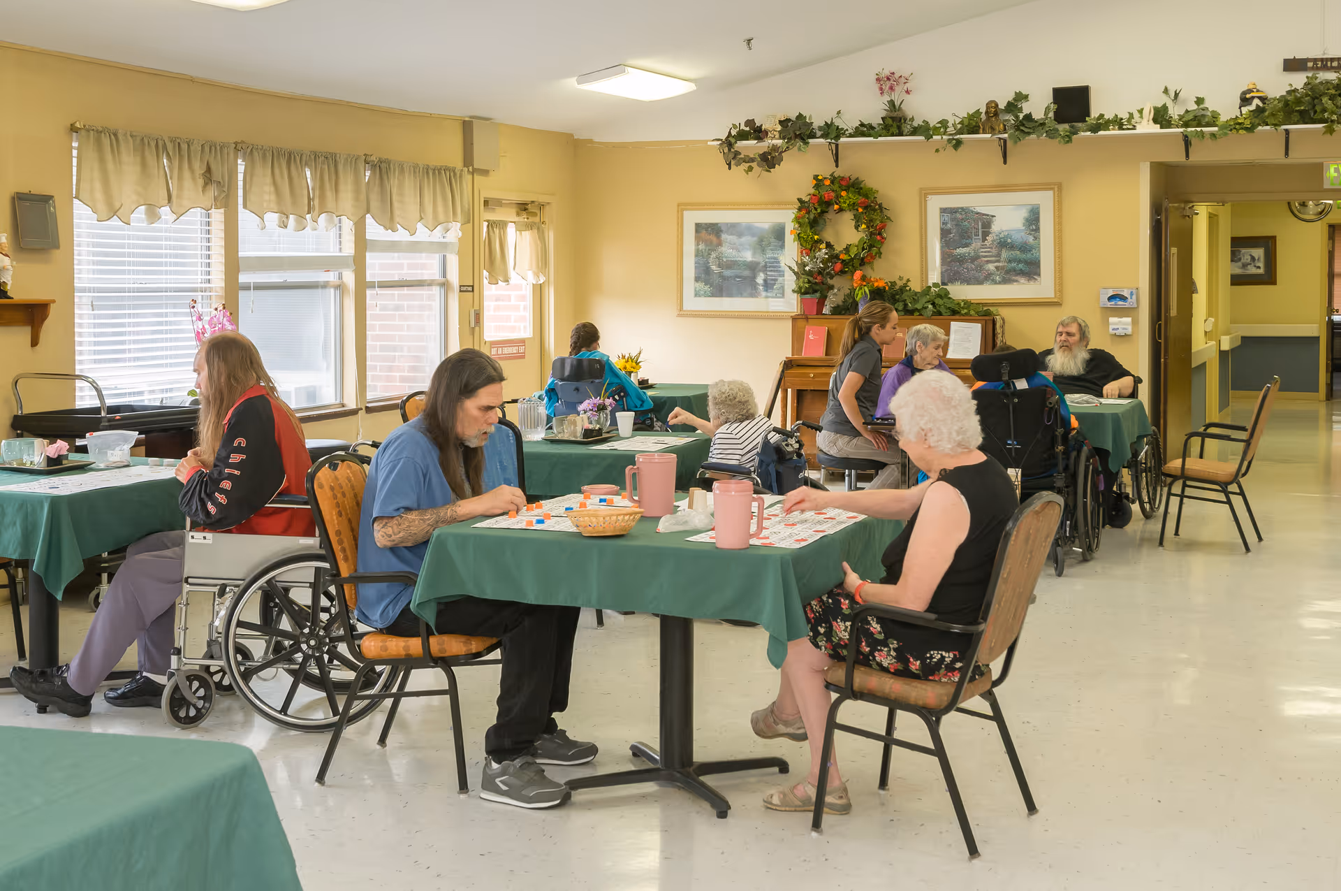 A group of elderly people and a caregiver in a common room of a care center. Several individuals are seated at tables covered with green tablecloths, engaging in activities such as playing games. Some people are in wheelchairs. The room has large windows with curtains, framed pictures on the walls, and decorative plants.