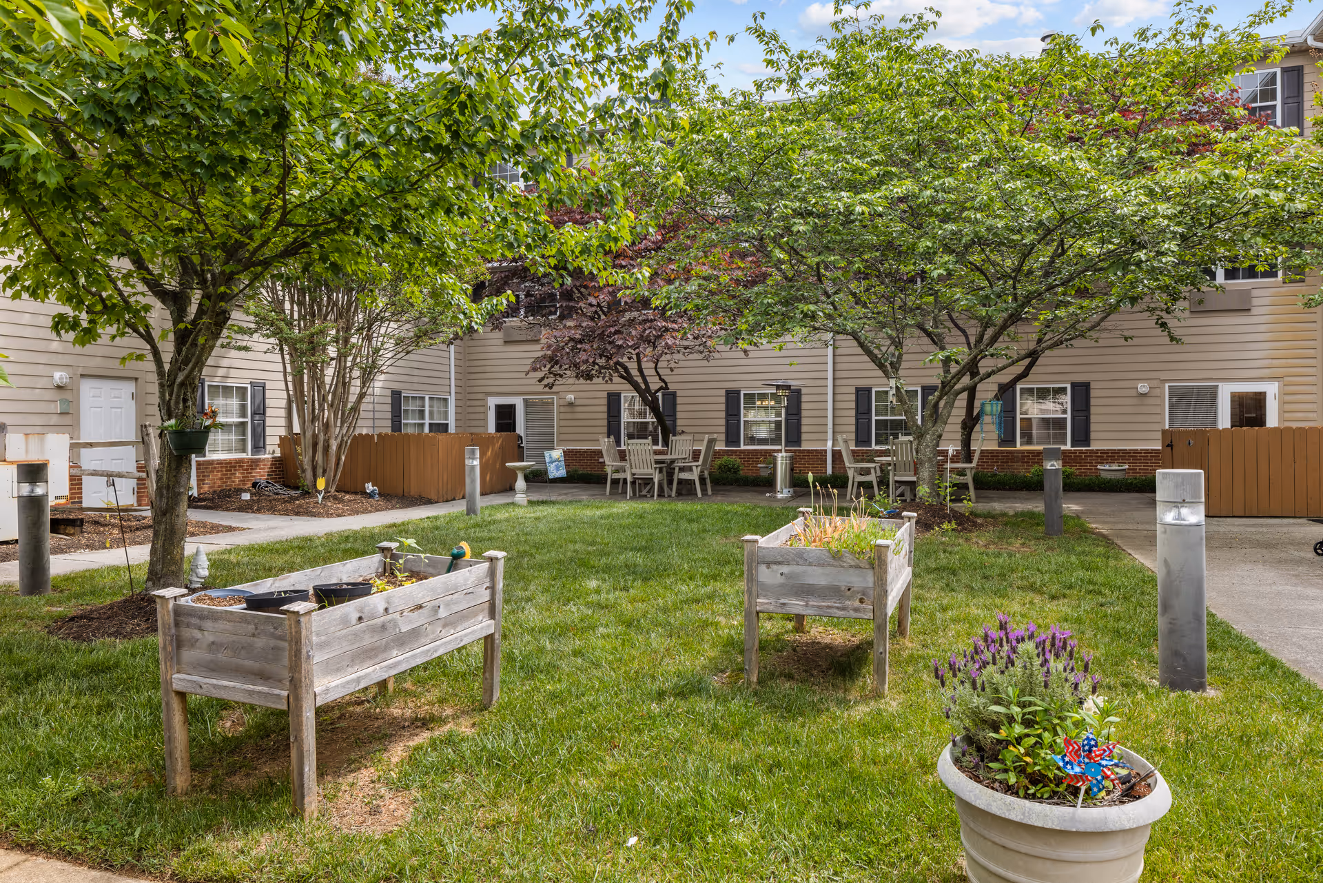 A grassy courtyard with raised wooden planters, potted flowers, trees and patio seating in front of beige apartment buildings.