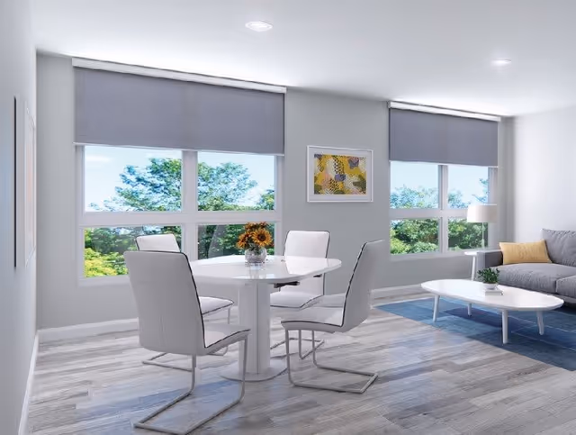 A bright and modern living area with large windows showing green trees outside. The room features a white round dining table with four white chairs, a gray sofa with a yellow pillow, a white coffee table on a blue rug, and a framed abstract artwork on the wall. The floor has light wood-style flooring and the windows have gray roller blinds.