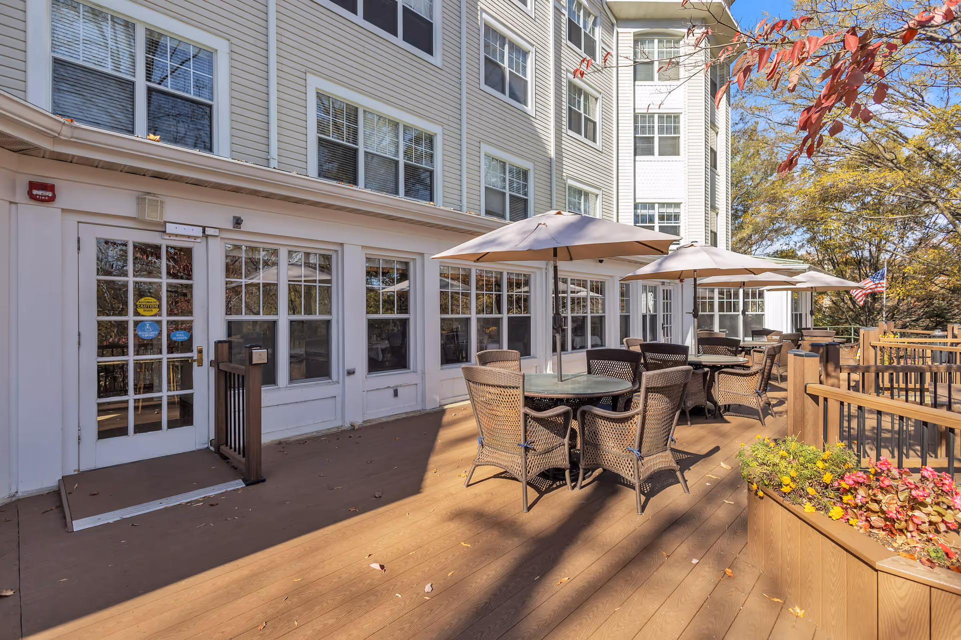 Outdoor patio area at Sunrise at Bluemont Park featuring multiple round tables with wicker chairs and large beige umbrellas. The patio is attached to a multi-story building with many windows. There are flower planters with colorful flowers and trees with autumn foliage surrounding the area.