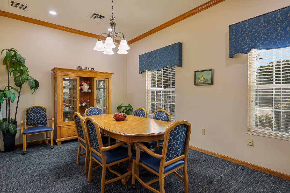 Wooden dining table with six blue-upholstered chairs in a well-lit common room with a china cabinet and a 'Welcome Home' sign.