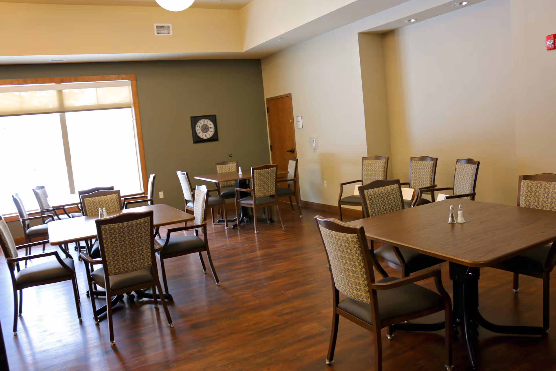 Sunlit dining room with several wooden tables and patterned upholstered chairs on hardwood floors.