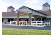 Exterior view of a single-story brick building with a covered entrance supported by four stone columns, white fencing, and two cupolas on the roof, set against a clear sky with a grassy lawn in the foreground.