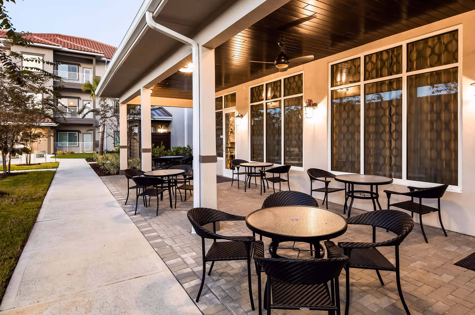 Outdoor covered patio area at Windsor Pointe of Jacksonville with several round tables and black wicker chairs. The patio has a wooden ceiling with ceiling fans and wall-mounted lights. A concrete walkway runs alongside the patio, leading to a multi-story building with balconies and red-tiled roofs. There are some trees and landscaped greenery around the walkway.