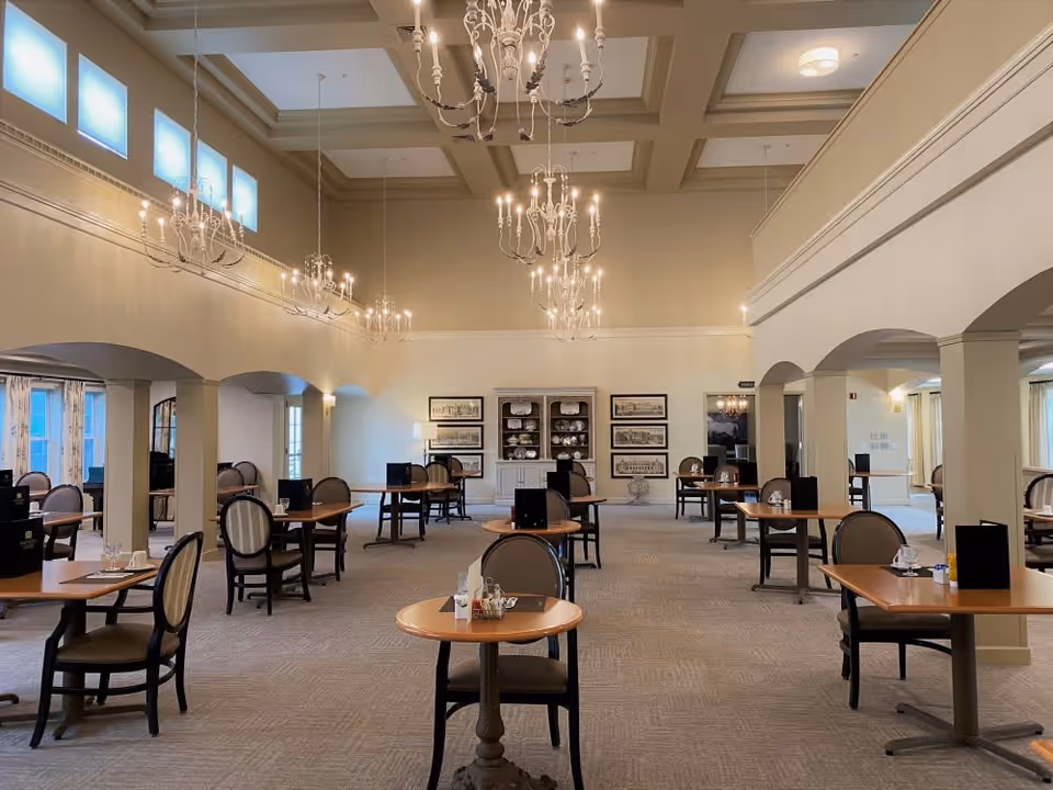 A spacious dining room in a senior living facility with multiple wooden tables and cushioned chairs arranged neatly. The room features high ceilings with elegant chandeliers, large windows allowing natural light, and decorative framed pictures on the far wall. The atmosphere is calm and inviting.