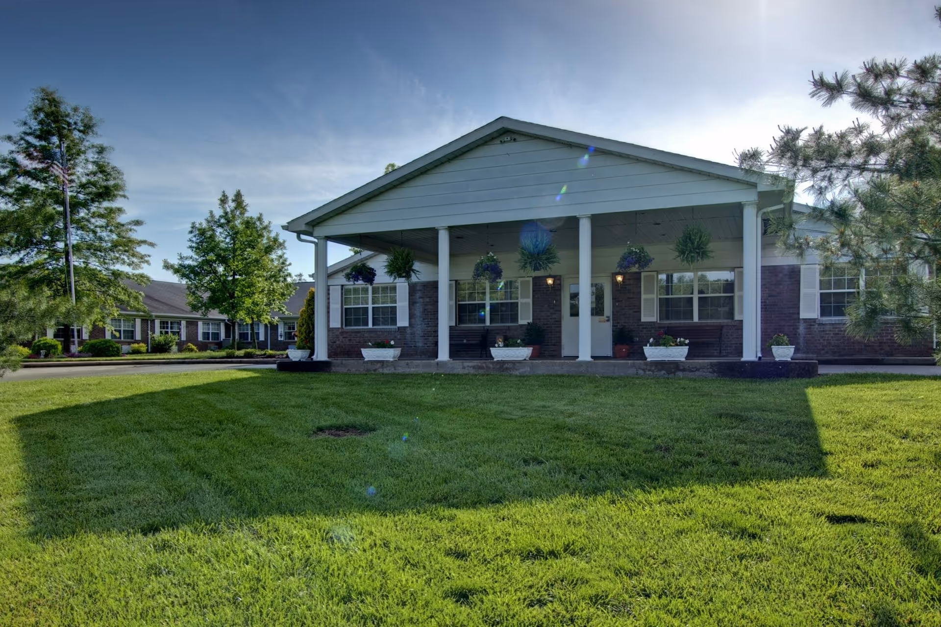 Front exterior view of a single-story brick building with white columns supporting a covered porch. The porch has hanging plants and flower pots along the edge. The building is surrounded by a well-maintained green lawn and trees under a clear blue sky.