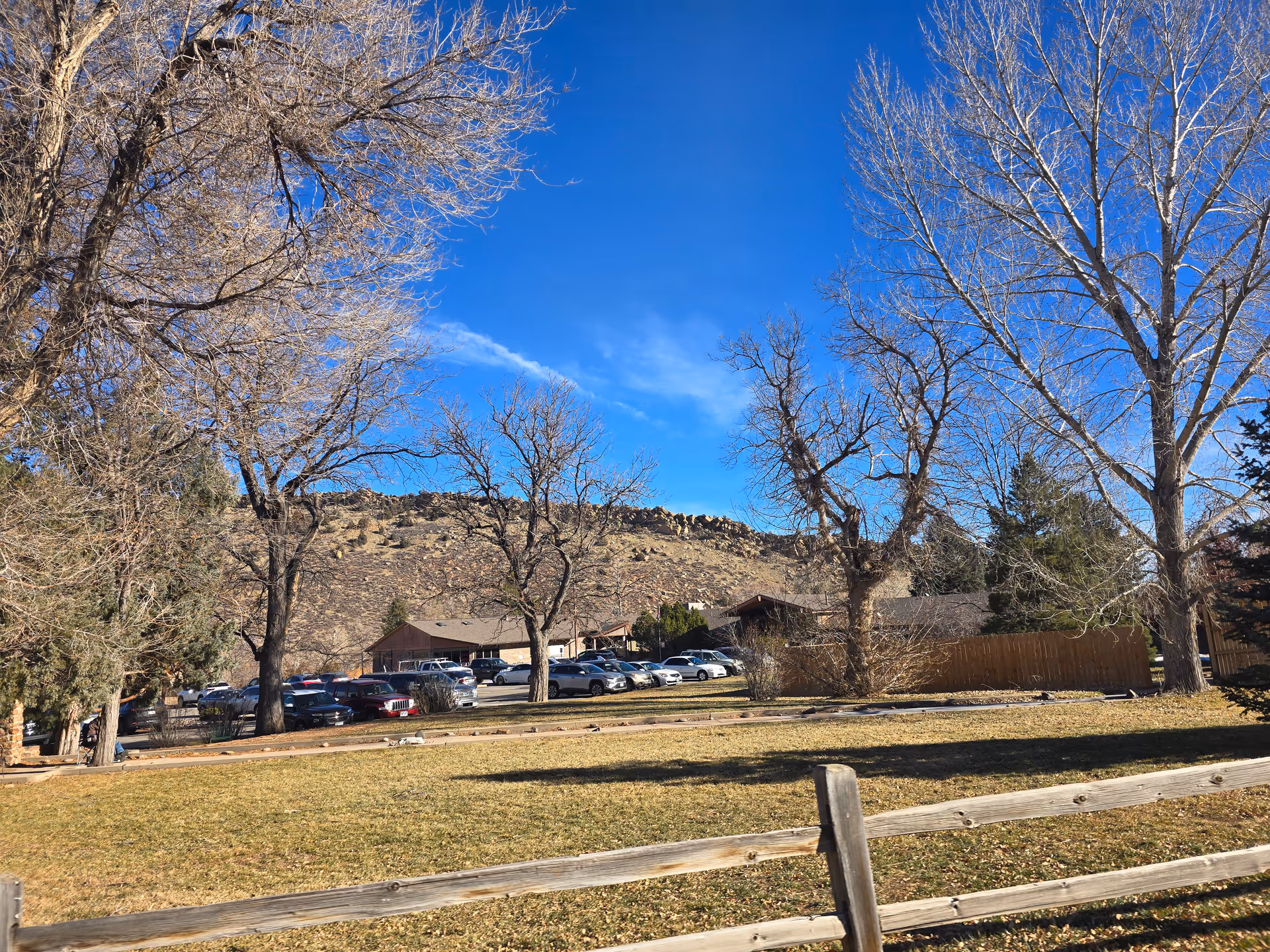 Outdoor view of Prestige Care Center of Morrison showing a grassy area with a wooden fence in the foreground, leafless trees, parked cars, and buildings with a rocky hill and clear blue sky in the background.