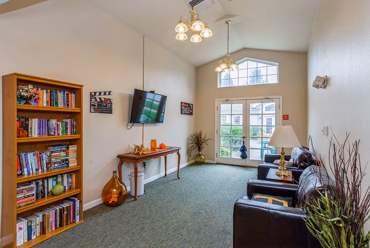 Bright communal living room with bookshelves, a wall-mounted TV, leather chairs and a table near glass doors to the outside.