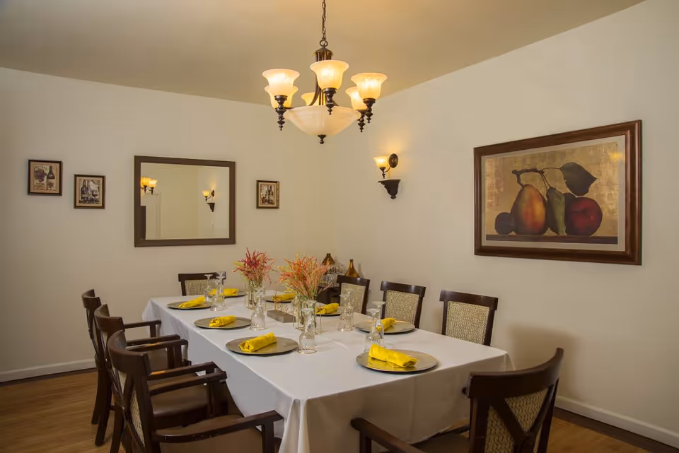 A dining room with a rectangular table covered with a white tablecloth, set with plates, glasses, and yellow napkins. The room has eight wooden chairs with cushioned seats around the table. There are two floral centerpieces on the table. The walls are decorated with a large framed painting of fruit, a mirror, and three smaller framed pictures. A chandelier with five lights hangs from the ceiling, and two wall sconces provide additional lighting.