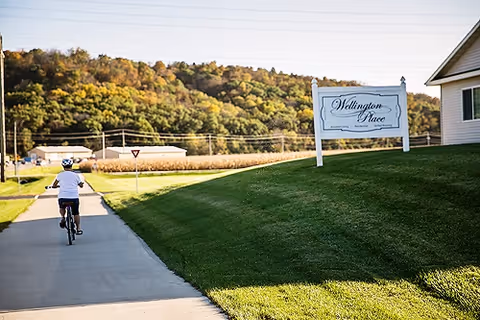 A person riding a bicycle on a paved path next to a grassy hill with a sign that reads 'Wellington Place'. In the background, there are trees with autumn foliage and some buildings.