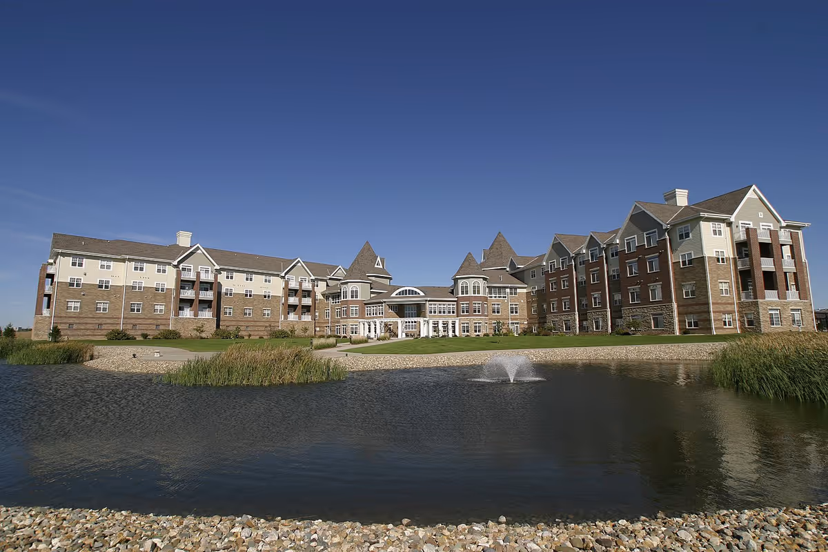 A large senior living facility building with multiple floors and peaked roofs, situated behind a pond with a small fountain. The building has a mix of brick and light-colored siding, with balconies and many windows. The sky is clear and blue.