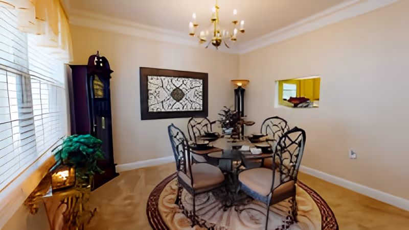 A dining room with a round glass table set for four, surrounded by four ornate metal chairs with cushioned seats. The room features beige walls, a decorative rug under the table, a tall grandfather clock in the corner, a chandelier hanging from the ceiling, and a small window opening to an adjacent room. There is also a plant on a small table near a large window with blinds.