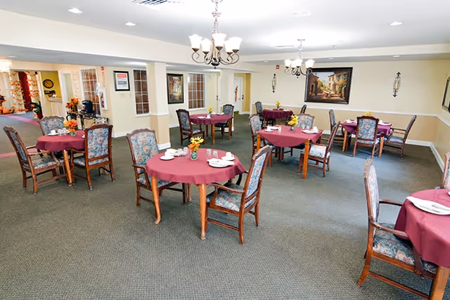A dining room with multiple round tables covered with burgundy tablecloths, each set with white plates, cups, and small floral centerpieces. The room has carpeted floors, beige walls with white trim, chandeliers hanging from the ceiling, and framed artwork on the walls.