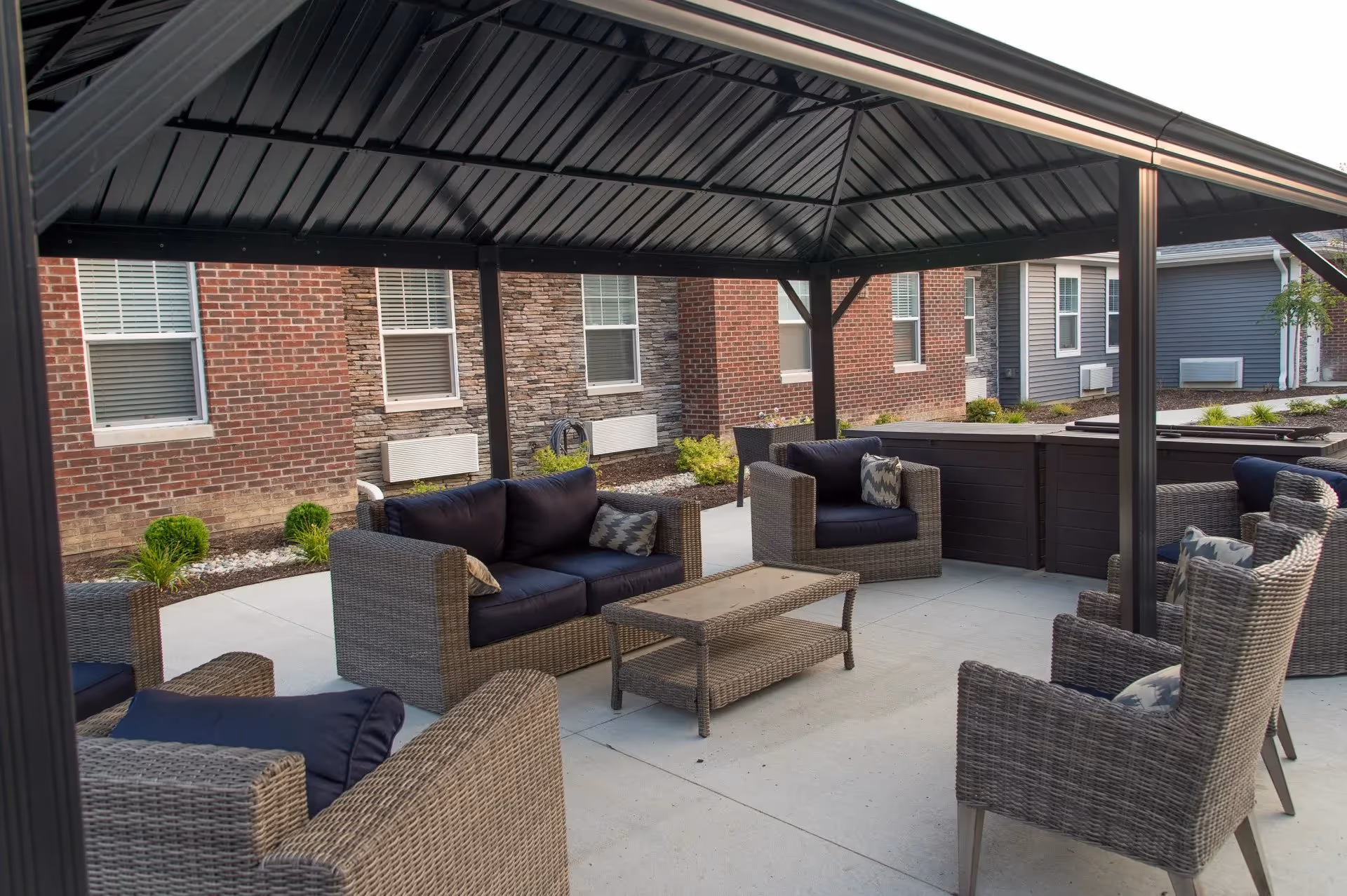 Outdoor covered seating area with wicker furniture including chairs and a loveseat with dark cushions and patterned pillows, set around a rectangular wicker coffee table. The area is paved with concrete and surrounded by brick and stone building walls with windows and some landscaping.
