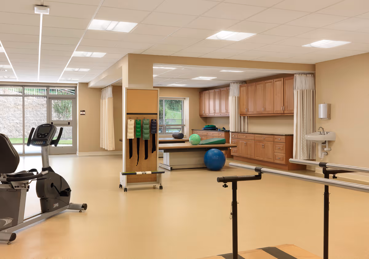 A spacious physical therapy or rehabilitation room with exercise equipment including a stationary bike, parallel bars, therapy balls, and a treatment table. The room has beige walls, large windows letting in natural light, wooden cabinets, and a wall-mounted sink with a soap dispenser.