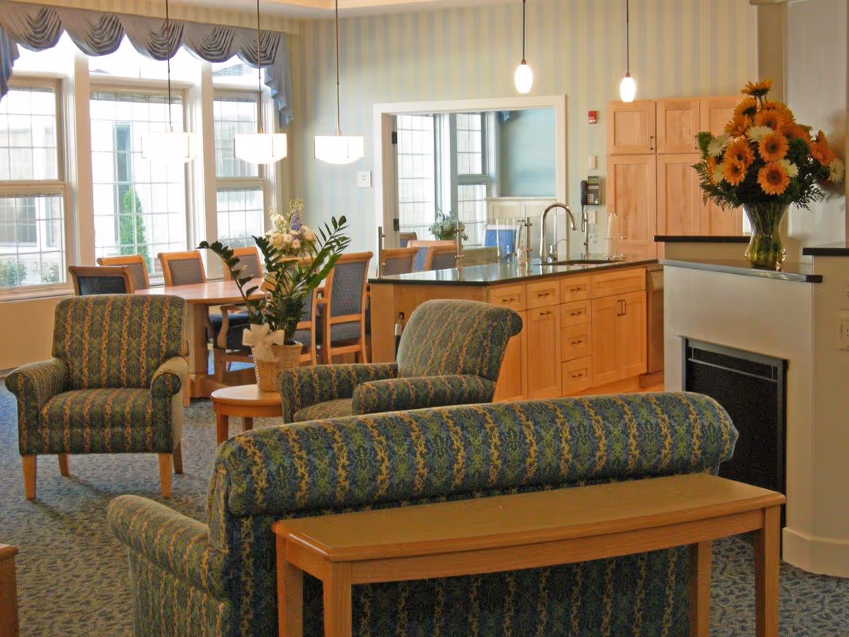 A cozy common area in a senior living facility featuring patterned upholstered armchairs and a sofa, a wooden coffee table with a potted plant, a kitchen island with a sink and wooden cabinetry, a dining table with chairs near large windows, and a vase of sunflowers on a counter next to a fireplace.