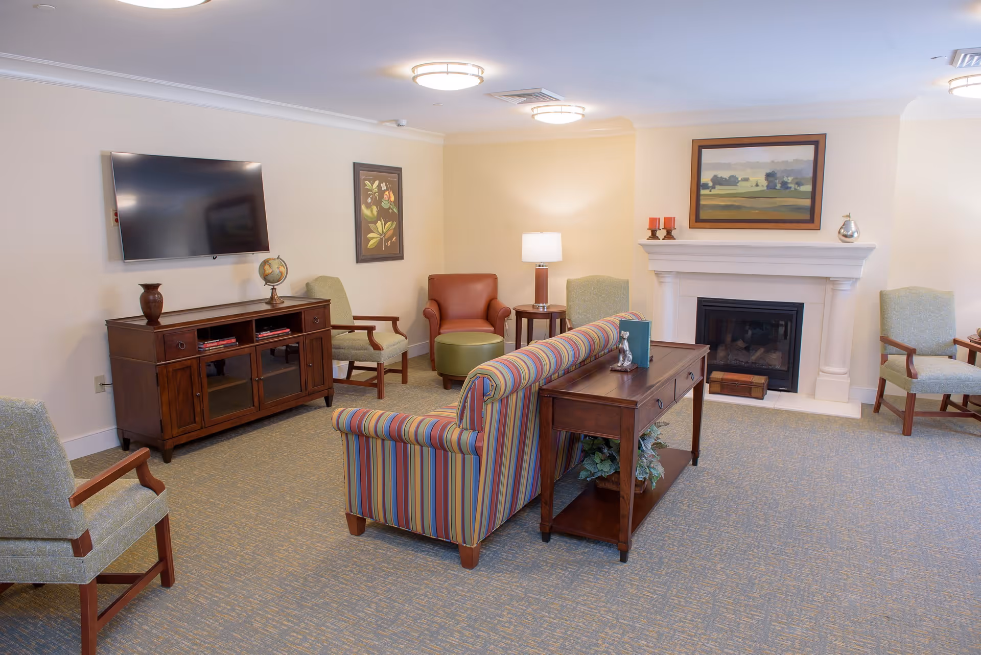A cozy living room with a striped sofa facing away, several armchairs, a wooden TV stand with a flat-screen TV mounted on the wall, a side table with a lamp, and a white fireplace with a landscape painting above it.