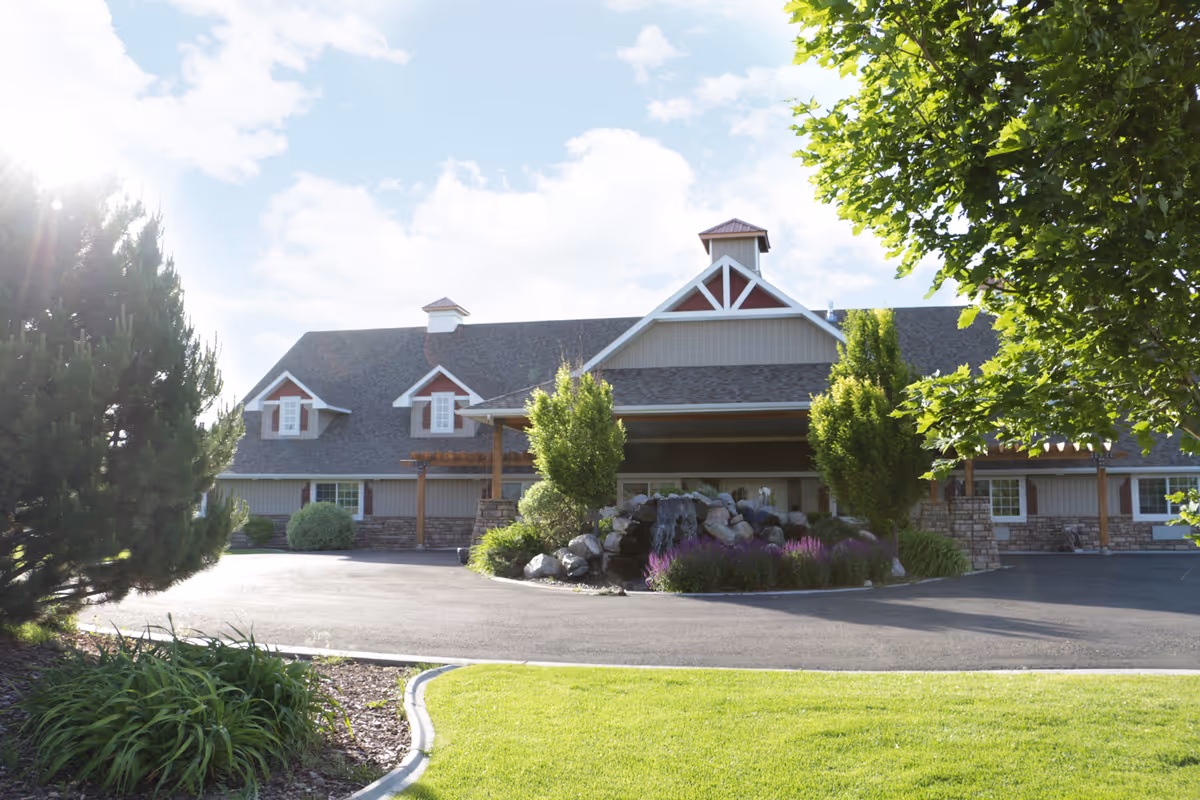 Exterior view of a large building with a peaked roof, surrounded by trees and landscaping including a rock garden with purple flowers. The sky is partly cloudy with sunlight shining through.