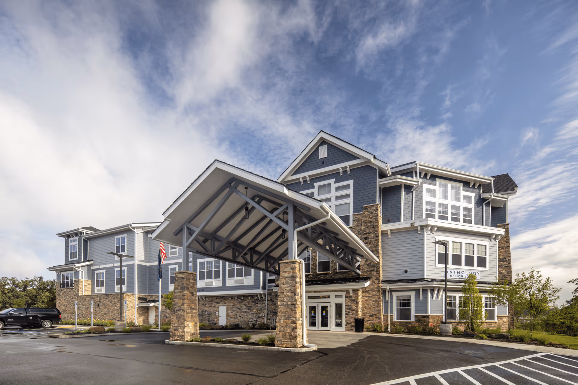 Exterior view of a modern senior living facility building with a covered entrance supported by stone pillars. The building has blue and gray siding with stone accents and multiple large windows under a partly cloudy sky.