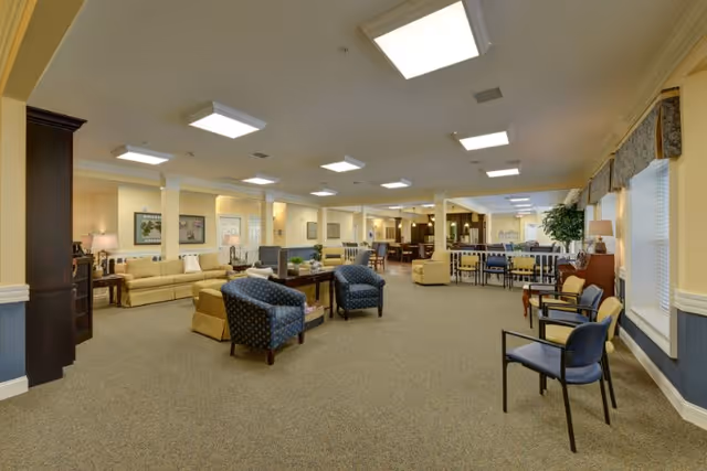 A spacious and well-lit common area in a senior living facility featuring multiple seating arrangements including sofas, armchairs, and dining tables. The room has beige walls with white trim, carpeted floors, large windows with valances, and ceiling lights providing ample illumination.