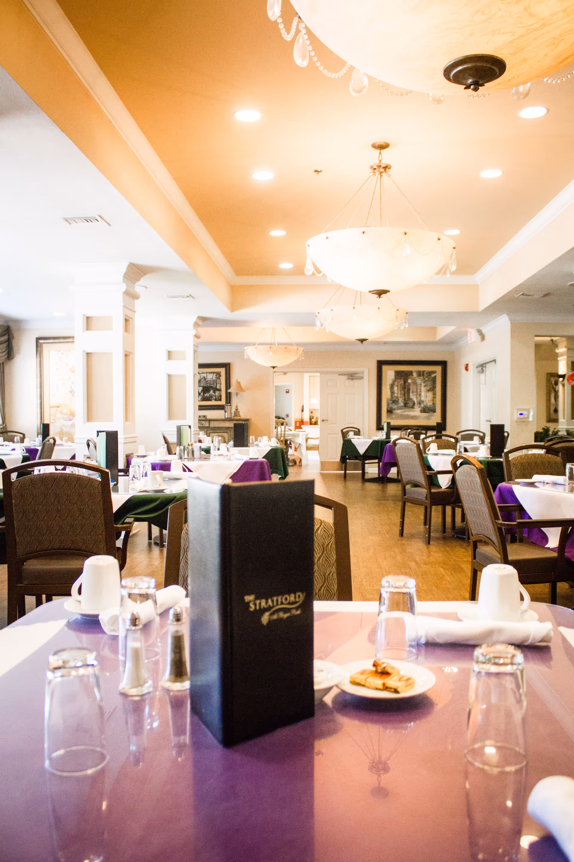 Interior view of a dining room in a senior living facility with tables covered in purple and green tablecloths. Each table is set with glasses, cups, napkins, and silverware. A black menu with 'The Stratford' written on it is prominently placed on the nearest table. The room is well-lit with large chandeliers hanging from the ceiling and framed artwork on the walls.
