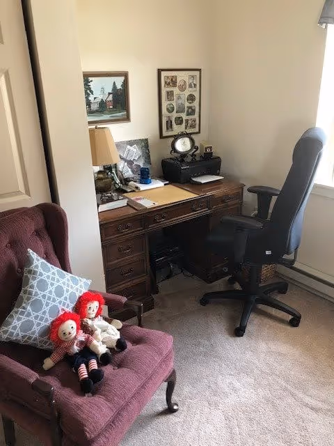 A cozy corner of a room featuring a wooden desk with various items including a printer, clock, and papers. Next to the desk is a black office chair. In the foreground, there is a maroon upholstered armchair with a patterned pillow and two Raggedy Ann dolls sitting on it. The walls have framed pictures, and a window with blinds allows natural light into the room.