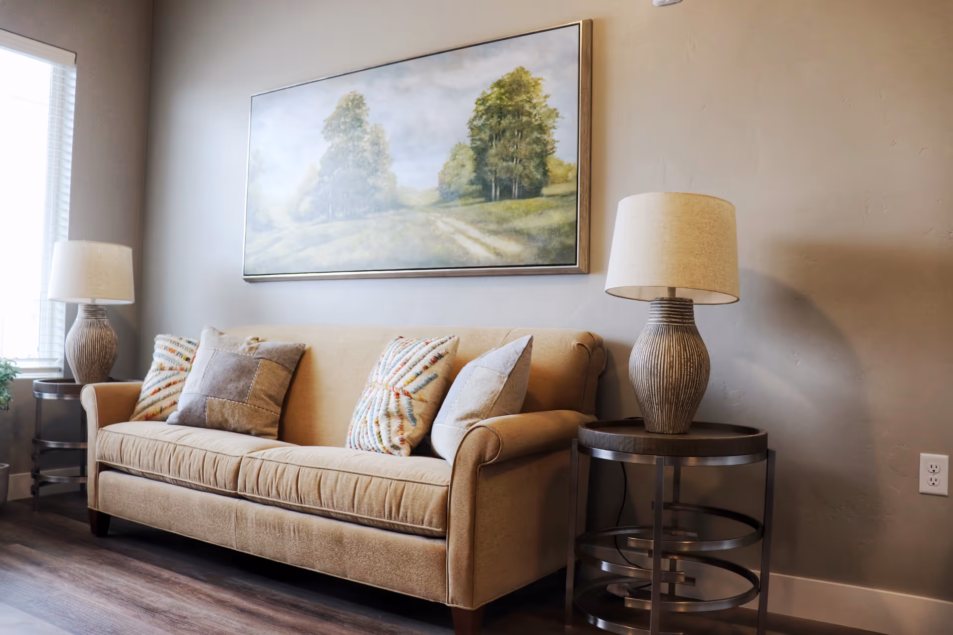 Beige sofa with patterned throw pillows between two side tables and lamps beneath a framed landscape painting.