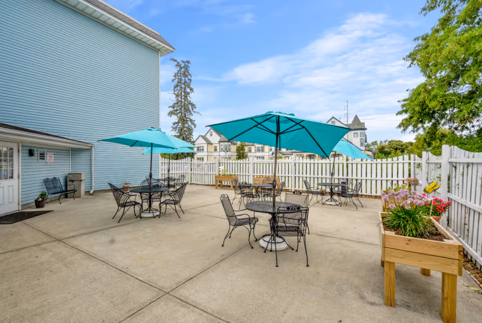 Outdoor patio area with several black metal tables and chairs, each table shaded by a teal umbrella. The patio is surrounded by a white picket fence and has raised wooden planters with flowers. A light blue building is visible on the left side, and trees and other buildings can be seen in the background under a partly cloudy sky.