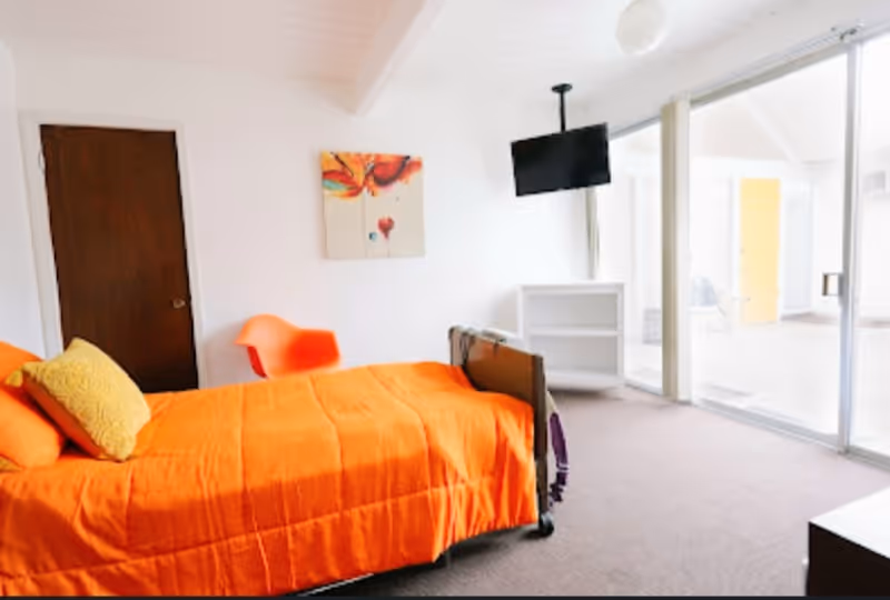 Bright bedroom with orange bedspread, mounted TV, and glass sliding doors in a senior facility.