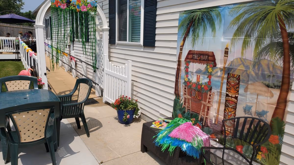 Outdoor patio area at Timberwood Lodge Assisted Living with plastic chairs and tables, a white fence, a decorative archway with hanging green vines and flowers, a potted plant, and a tropical-themed backdrop featuring palm trees, a tiki bar, and tiki totems.