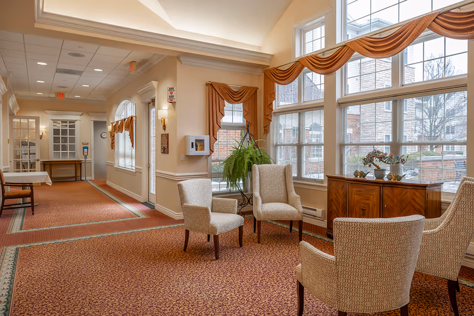 Sunlit common seating area with upholstered chairs, large windows, and a carpeted hallway.